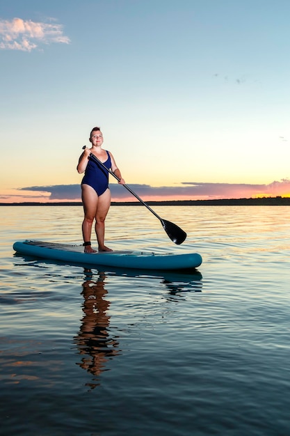 A Jewish feminist woman in a closed swimsuit with a mohawk standing on a SUP board with an oar floats on the water against the background of the sunset sky