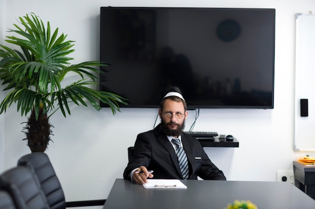 Jewish man in a yarmulke and glasses is sitting at a table in office