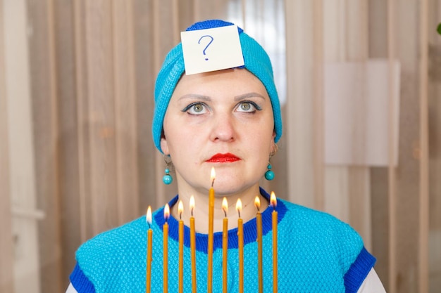 A Jewish woman in a kisui rosh headdress with a Hanukkah candlestick with burning candles in her hands is thinking whether she has lit the candles correctly. Horizontal photo