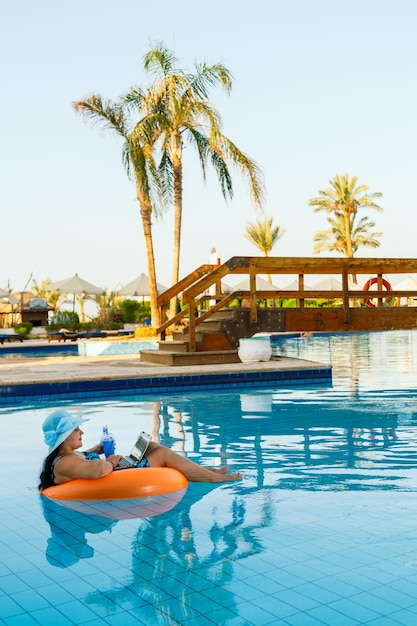 A Jewish woman in a sun hat in a swimming pool in a swimming circle with a cocktail and a laptop communicates via video link on the background of palm trees.