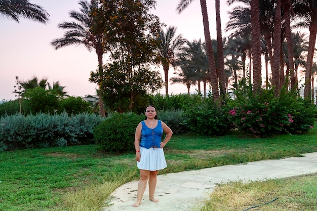 A Jewish woman in a white skirt walks along a path near the palm trees and the sea in the evening at sunset. Horizontal photo