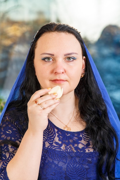 A Jewish woman with her head covered in a blue cape at the Passover Seder table performs the commandment karpas with a bow. Vertical photo