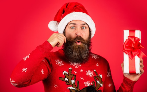 Jingle Bells. winter vakantie feest. volwassen man viert nieuwjaar. bebaarde hipster in kersttrui. verraste man met baard in Kerstman hoed. brutale man houdt de huidige doos vast. kerst online winkelen.