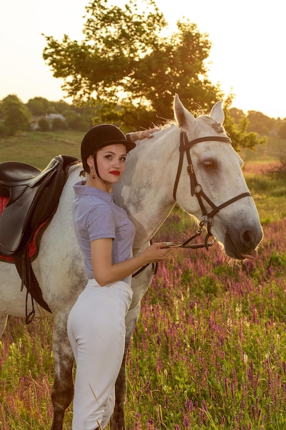 Jockey young girl petting and hugging white horse in evening sunset. Sun flare