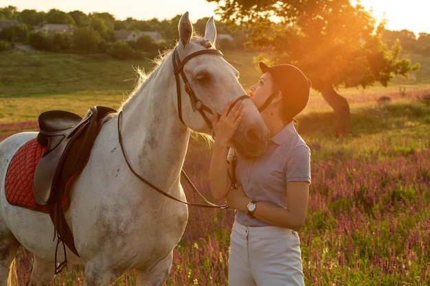 Jockey young girl petting and hugging white horse in evening sunset. Sun flare
