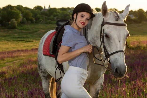 Jockey young girl petting and hugging white horse in evening sunset. Sun flare