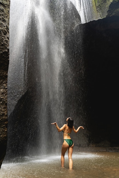 Join me. Kind brunette girl wearing swimsuit and standing in front of waterfall, wild nature