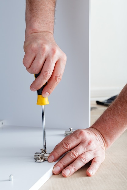 Joiner verzamelt meubels met behulp van handgereedschap schroevendraaier. meubelmontage met behulp van een schroevendraaier. verhuizen, woningverbetering, meubelreparatie en renovatie. timmerman verzamelt een witte meubelkast.