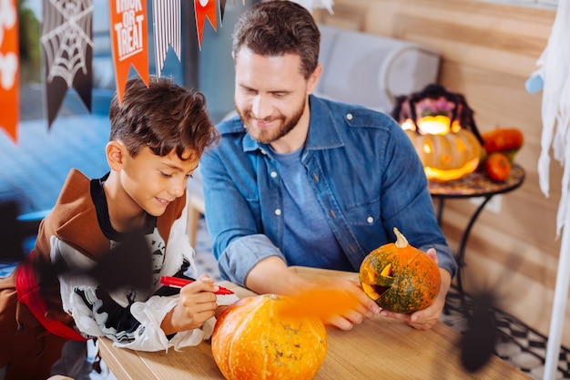 息子に加わります。家族と一緒に自宅でハロウィーンパーティーのためにカボチャを着色彼のかわいい息子に参加する愛情のあるひげを生やした父