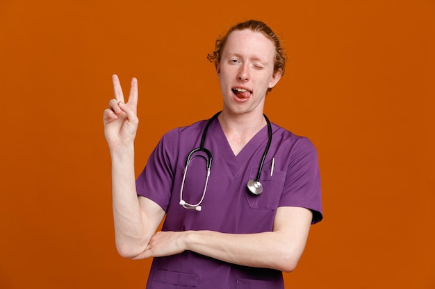 Jokin showing tongue with peace gesture young male doctor wearing uniform with stethoscope isolated on orange background