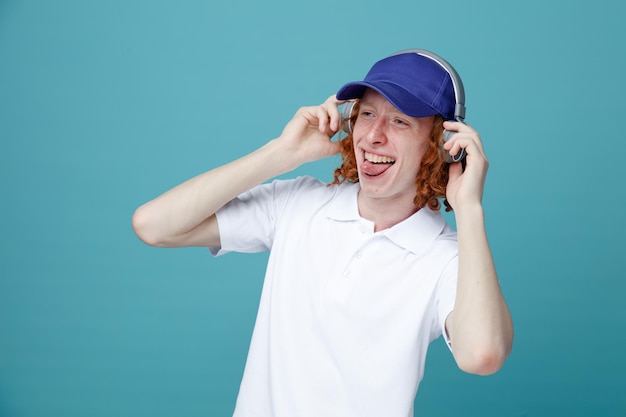 Jokin showing tongue young handsome guy in cap wearing headphones isolated on blue background