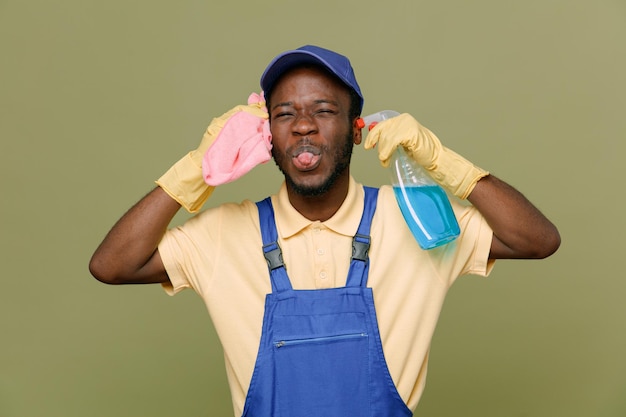 Шутка показывает язык, держащий чистящее средство с тряпкой, молодой афроамериканец-уборщик в униформе с перчатками, изолированными на зеленом фоне