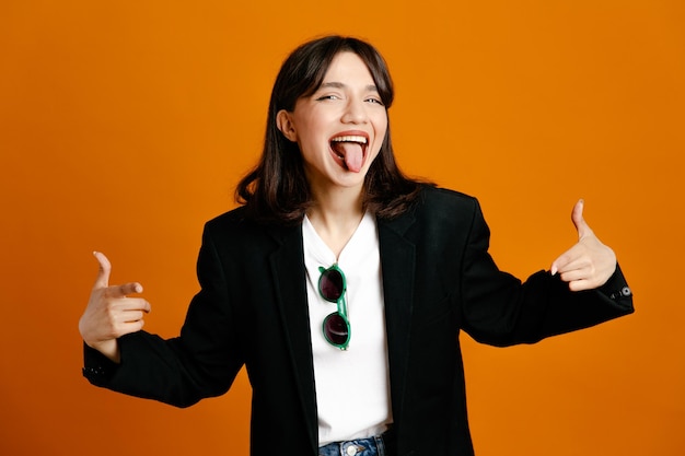 Joking showing tongue young beautiful female wearing black jacket isolated on orange background