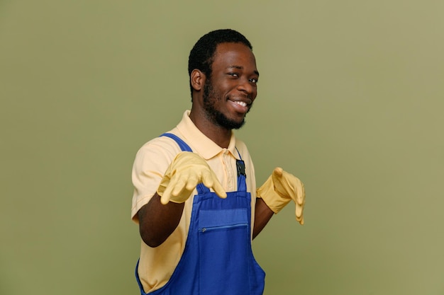 Joking young africanamerican cleaner male in uniform with gloves isolated on green background