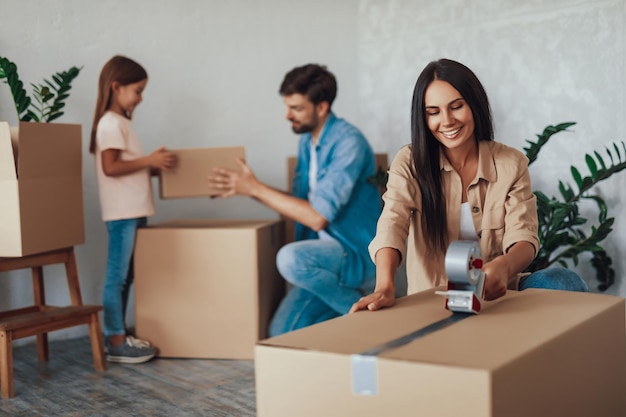 Jolly brunette lady taping up cardboard boxes while her husband and daughter moving boxes on the background