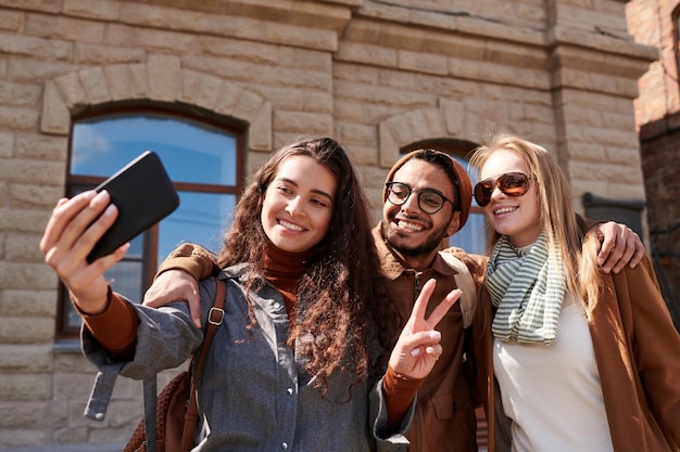 Jolly young multiethnic friends embracing each other and posing for selfie against old building