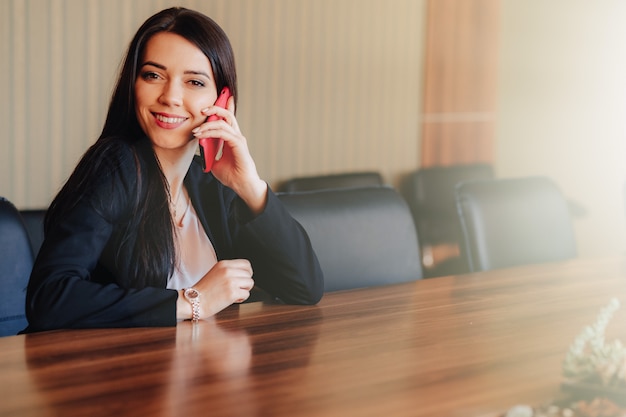 Jong aantrekkelijk emotioneel meisje in bedrijfsstijlkleren die bij bureau met telefoon zitten