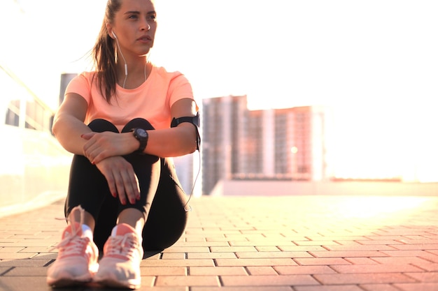 Jong aantrekkelijk sportief meisje dat op straat rust met mobiele telefoon en muziek, buiten bij zonsondergang of zonsopgang in de stad