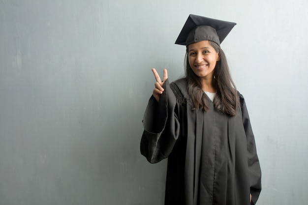 Jong afgestudeerd Indiase vrouw tegen een muur plezier en gelukkig, positief en natuurlijk, het doen van een gebaar van de overwinning, vrede concept