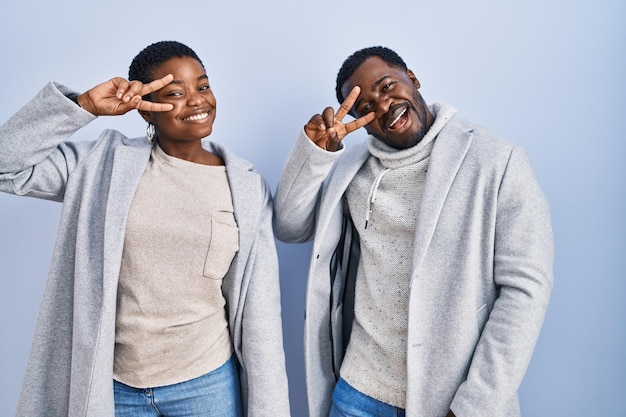 Jong afrikaans amerikaans paar dat zich over blauwe achtergrond bevindt die samen vredessymbool doen met vingers over gezicht glimlachend vrolijk tonend overwinning