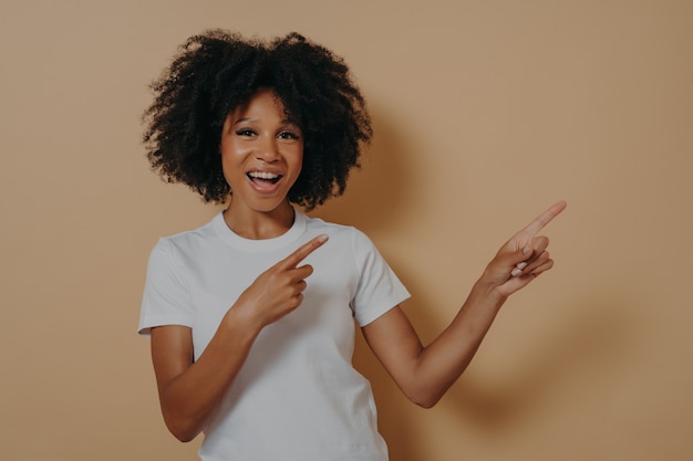 Jong afro-amerikaans meisje draagt casual kleding vrolijk en breed glimlachend terwijl ze met beide handen en wijsvingers naar de zijkant wijst met enthousiaste uitdrukking op haar gezicht, kopieer ruimte