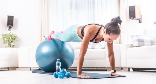 Jong atletisch meisje houdt een plankpositie in haar woonkamer met benen bovenop de fitnessbal