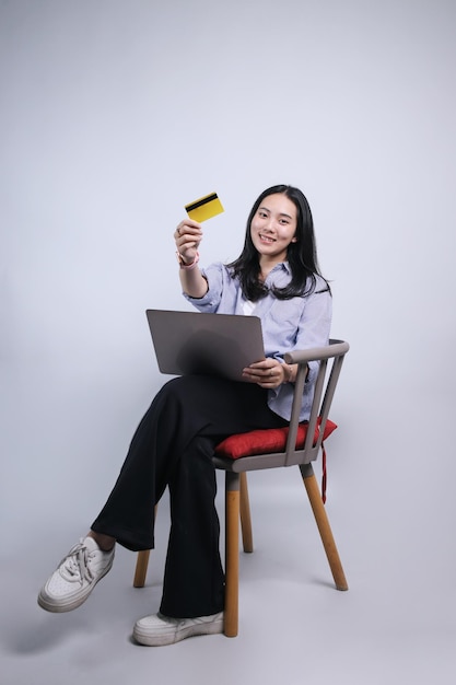 Jong Aziatisch meisje toont een creditcard terwijl ze op een stoel zit met een laptop op haar schoot