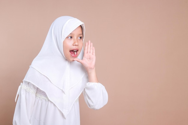 Jong Aziatisch moslimmeisje draagt een hijab, legt haar hand bij haar mond en schreeuwt of roept iemand.