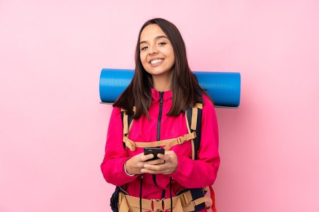 Jong bergbeklimmermeisje met een grote rugzak over geïsoleerd roze die een bericht met mobiel verzenden