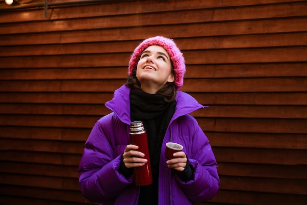 Jong blij dat een vrouw hete thee drinkt terwijl ze een thermoskan vasthoudt op een winterse dag om op te warmen in...