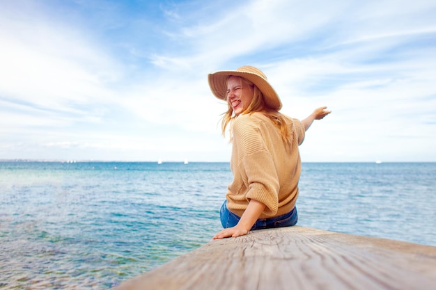Jong blij meisje in een zonnehoed zit op een pier aan zee en verheugt zich over reizen en vrijheidsconcept
