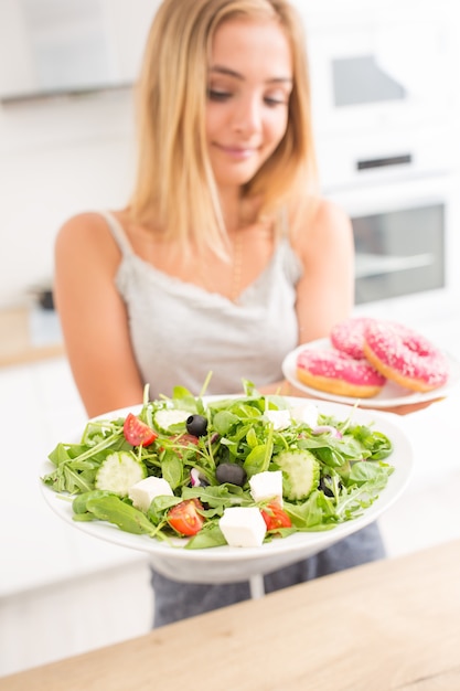 Jong blond meisje bij het ontbijt of diner in de keuken thuis kiest tussen donut en groentesalade.