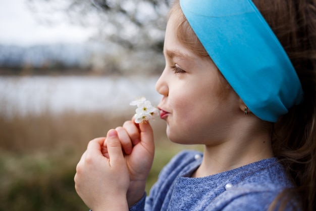 Jong blond meisje dat zich in een bloeiende tuin bevindt. Bloeiende kers. Portret van mooi meisje. Sluit omhoog van klein modelgezicht. Sakura bloeiend, lenteavondtijd