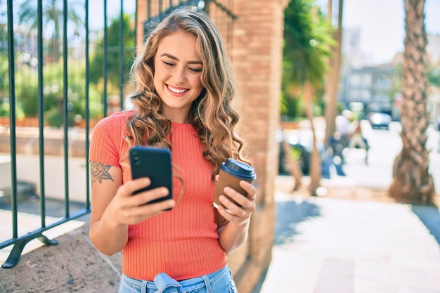 Jong blond meisje glimlachend gelukkig met behulp van smartphone en het drinken van afhaalkoffie in de stad.