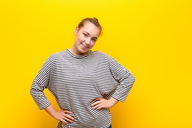 Jong blonde mooi meisje die gelukkig, vrolijk en zeker kijken, trots glimlachen en aan kant met beide handen op heupen tegen gele muur kijken