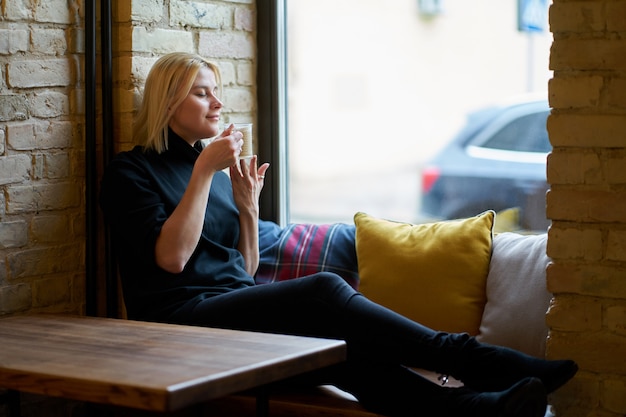 Jong blondemeisje dat dichtbij venster situeert en koffie in het koffie drinkt. Tijd om te ontspannen.