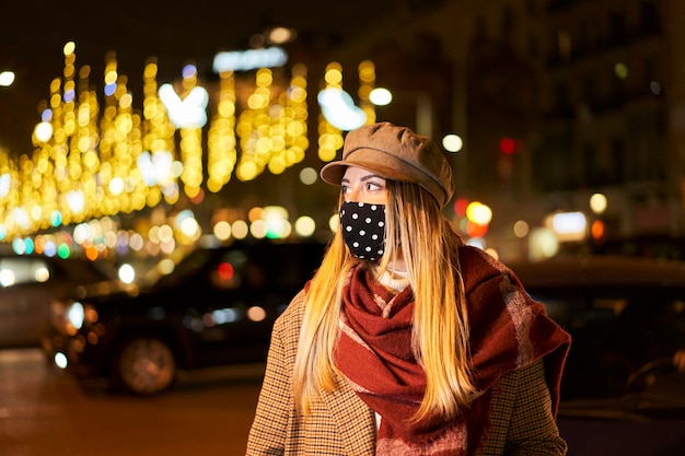 Jong blondemeisje die met masker aan de kant kijken. Ze is 's nachts in een stad, met veel achterlichten en passerende auto's. Winterse sfeer.