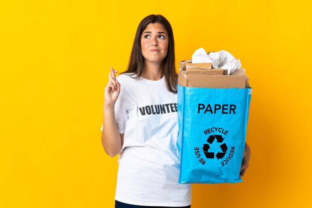 Jong Braziliaans meisje dat een recyclingzak vol papier houdt om te recyclen geïsoleerd op geel met vingers die kruisen en het beste wensen