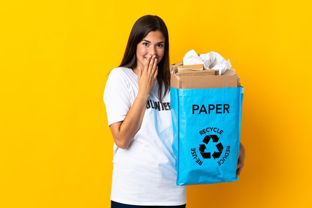 Jong Braziliaans meisje met een recyclingzak vol papier om te recyclen geïsoleerd op gele achtergrond gelukkig en glimlachend mond bedekken met hand