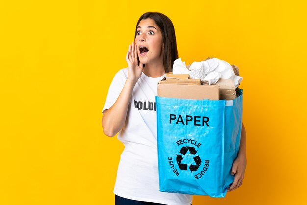 Jong Braziliaans meisje met een recyclingzak vol papier om te recyclen geïsoleerd op gele muur schreeuwen met mond wijd open naar de zijkant