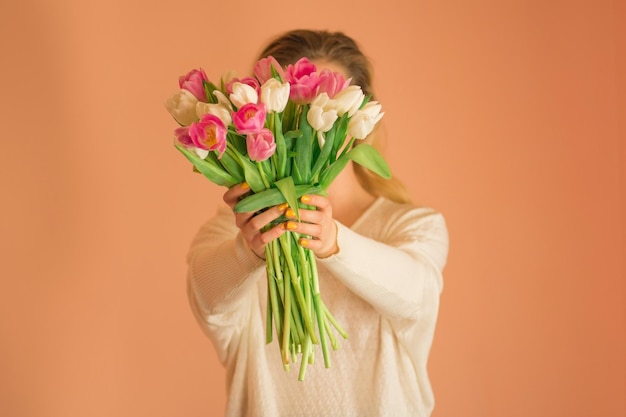 Jong charmant glimlachmeisje dat groot boeket tulpen op lichtbeige achtergrond houdt jong mooi meisje in de witte jurk met kleurrijke verse bloementulpen