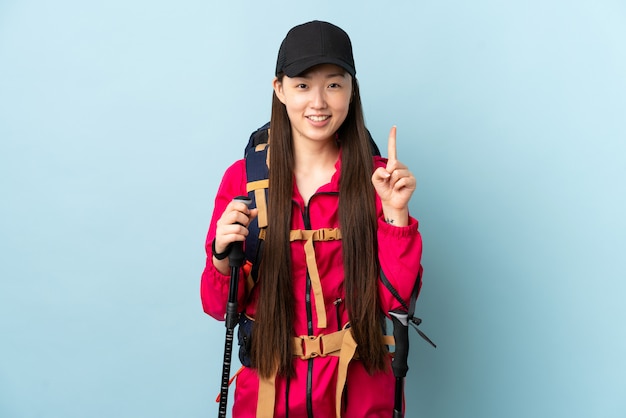 Jong Chinees meisje met rugzak en trekkingstokken over geïsoleerde blauwe muur die een groot idee benadrukken