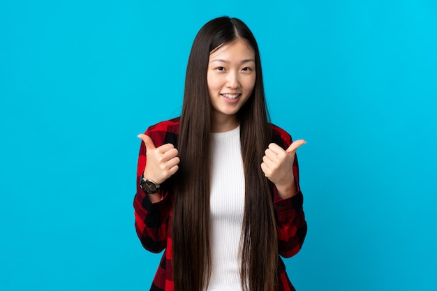 Jong Chinees meisje op geïsoleerd blauw met duimen op gebaar en het glimlachen