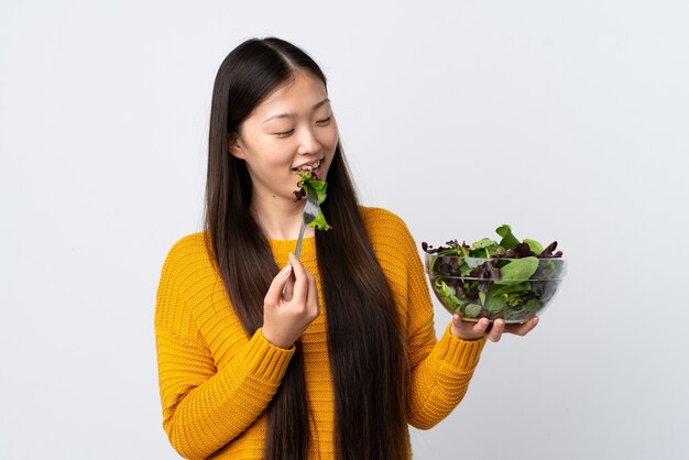 Jong Chinees meisje op geïsoleerd wit die een kom salade houdt en het met gelukkige uitdrukking bekijkt