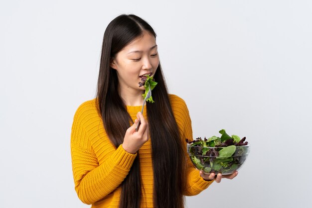 Jong Chinees meisje over geïsoleerde witte muur die een kom salade houdt en het met gelukkige uitdrukking bekijkt