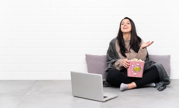 Jong Colombiaans meisje dat een kom popcorns houdt en een film toont in laptop die veel glimlacht