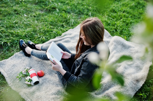 Jong donkerbruin meisje die bij jeans op plaid tegen de boom van de de lentebloesem zitten en het boek lezen.