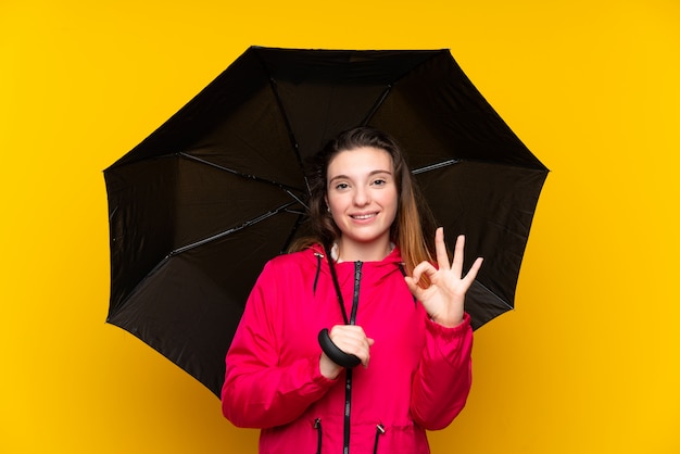 Jong donkerbruin meisje die een paraplu over geïsoleerde geel houden die ok teken met vingers tonen