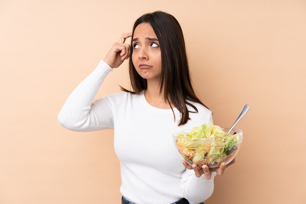 Jong donkerbruin meisje die een salade houden over het maken van het gebaar van waanzin die vinger op het hoofd zetten