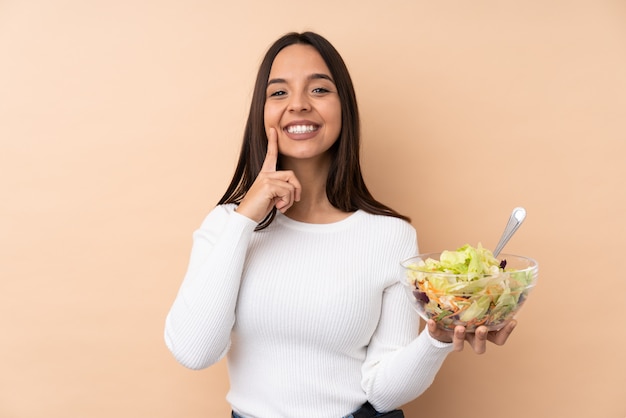 Jong donkerbruin meisje die een salade over geïsoleerde muur houden glimlachend met een gelukkige en prettige uitdrukking